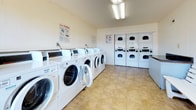 Located at 986 Armfield Circle in Norfolk, Virginia, this single-floor shared laundry facility is designed to serve the practical needs of its users with a clean, functional layout. The space is outfitted with a generous row of front-loading washing machines along one wall and a bank of stacked dryers along the opposite wall, providing ample capacity for multiple users simultaneously. Instructional signage is posted throughout to assist with machine operation. The flooring throughout is composed of neutral-toned tile, offering durability and ease of maintenance. White panel walls and overhead fluorescent lighting create a bright, well-lit environment. A white wooden bench is positioned along the wall, providing a comfortable waiting area for residents. A built-in counter surface is also available for folding or sorting laundry. Large glass doors and windows at one end of the room flood the space with natural light and offer a view of an enclosed outdoor courtyard, defined by brick walls and a railed upper walkway with mature trees visible beyond. The courtyard is accessible directly from the laundry room, adding a pleasant sense of openness to the otherwise utilitarian space. Overall, the facility is well-maintained and thoughtfully laid out to accommodate everyday laundry needs efficiently.