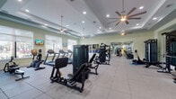 Located in Miramar, FL, this well-appointed fitness center offers a comprehensive workout environment spread across a single floor. The entry corridor sets an inviting tone with light wood-look tile flooring, a striking geometric linear chandelier, bold wall art, and a tall potted palm — creating a polished transition into the main facility.

The expansive main workout floor is defined by tray ceilings with recessed lighting and large ceiling fans that maintain comfortable airflow throughout. Rubber-tiled flooring provides safety and durability across the exercise zones. Multiple strength machines, cable systems, a Smith machine, and dumbbell racks with an adjustable bench cater to a full range of resistance training. Treadmills, elliptical trainers, and other cardio equipment are positioned near windows that invite natural light and outdoor views, each paired with mounted flat-screen TVs for entertainment.

A dedicated cycling studio features a row of stationary spin bikes alongside wall mirrors and motivational artwork, offering an immersive group fitness experience. Full-length mirrors throughout the facility amplify the sense of openness. Additional amenities include a dual water fountain and bottle-fill station conveniently located near a utility corridor. The overall ambiance is clean, bright, and energetic, reflecting a facility designed to support a full spectrum of fitness goals.
