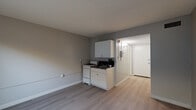 Located at 15001 Crenshaw Blvd in Gardena, California, this single-floor unit offers a practical and well-organized layout across four distinct spaces: a living room, bathroom, hallway, and closet. The living room serves as the primary multipurpose area, featuring light-toned hardwood-style flooring, neutral gray walls, and a wall-mounted air conditioning unit beneath a window that allows natural light to filter in. A compact kitchen is integrated into the living area, equipped with white cabinetry, a gas range, and a sink with dark countertops — providing efficient functionality within a modest footprint. The hallway connects the main living space to the entry door and features a textured ceiling, wall sconce lighting, and a sliding closet door for convenient storage access. A utility panel is also visible along the hallway wall. The bathroom is finished with floor-to-ceiling dark earth-toned tile surrounding a full soaking tub and shower combination, complemented by a white vanity and toilet. The tile detailing adds a bold, contrasting design element to the otherwise light and understated palette of the unit. Throughout the space, clean white baseboards and consistent flooring tie the rooms together cohesively, creating a functional and straightforward living environment.