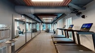 Located at 55 Romney St in Charleston, South Carolina, this well-appointed fitness center offers a thoughtfully designed single-floor layout that accommodates a full range of workout activities. The space is divided into two distinct functional zones, each purpose-built to support different training disciplines.

The cardio area lines one side of the facility and features a row of modern treadmills and upright bikes, each equipped with interactive digital displays. Large windows along the exterior walls flood this zone with natural light and provide views of the surrounding neighborhood, creating an energizing workout environment.

A floor-to-ceiling glass partition wall elegantly separates the cardio zone from the strength training area, maintaining an open, connected feel throughout the facility. The strength room is outfitted with a power rack, adjustable benches, medicine balls, and a variety of free weights, all arranged on durable rubberized flooring designed for heavy use.

The design aesthetic is distinctly industrial-modern: exposed ductwork and large cylindrical HVAC conduits run overhead against a dark ceiling, contrasted beautifully by warm wood-slat ceiling panels that add texture and visual warmth. Linear LED lighting throughout ensures excellent visibility across all areas. A separate spin studio with glass entry doors rounds out the facility, offering a dedicated space for cycling classes. The overall ambiance is sleek, functional, and motivating.