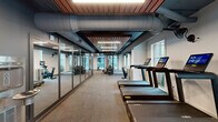 Located at 55 Romney St in Charleston, South Carolina, this well-appointed fitness center offers a thoughtfully designed single-floor layout that accommodates a full range of workout activities. The space is divided into two distinct functional zones, each purpose-built to support different training disciplines.

The cardio area lines one side of the facility and features a row of modern treadmills and upright bikes, each equipped with interactive digital displays. Large windows along the exterior walls flood this zone with natural light and provide views of the surrounding neighborhood, creating an energizing workout environment.

A floor-to-ceiling glass partition wall elegantly separates the cardio zone from the strength training area, maintaining an open, connected feel throughout the facility. The strength room is outfitted with a power rack, adjustable benches, medicine balls, and a variety of free weights, all arranged on durable rubberized flooring designed for heavy use.

The design aesthetic is distinctly industrial-modern: exposed ductwork and large cylindrical HVAC conduits run overhead against a dark ceiling, contrasted beautifully by warm wood-slat ceiling panels that add texture and visual warmth. Linear LED lighting throughout ensures excellent visibility across all areas. A separate spin studio with glass entry doors rounds out the facility, offering a dedicated space for cycling classes. The overall ambiance is sleek, functional, and motivating.