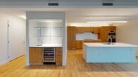 This contemporary single-floor residence offers a thoughtfully designed open-concept layout finished with warm light hardwood flooring throughout the main living areas. The expansive living space is defined by large black-framed windows on multiple walls, flooding the interior with natural light and framing striking panoramic city skyline views. A concrete structural column adds an industrial architectural accent to the open living area.

The kitchen is a standout feature, equipped with rich natural wood cabinetry, a sleek white quartz island with an integrated sink, a cooktop, double stainless steel wall ovens, and a built-in wine refrigerator. A wet bar area with glass shelving and an additional sink provides an elegant entertaining station adjacent to the main kitchen.

A wide hallway connects the living areas to the private rooms, with a plush-carpeted bedroom that opens directly to the kitchen and living spaces. The primary bathroom is finished with large-format dark stone-effect tiles, a deep soaking tub, a floating wood vanity with a white countertop, and a backlit vertical mirror. A separate water closet with neutral tile flooring provides added privacy and convenience. The overall aesthetic is clean, modern, and sophisticated, with a neutral palette throughout.