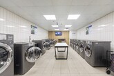 Located at 3634 Bathurst Street in Toronto, this commercial laundromat offers a clean, efficiently organized space designed to serve a high volume of customers. The single-floor layout is bright and welcoming, illuminated by recessed panel lighting and skylights embedded in a drop-tile ceiling that keeps the environment feel open and airy.

The main laundry area lines the walls with rows of modern stainless-steel front-load washing machines and large-capacity dryers, providing ample options for customers. A utility sink station is integrated within the machine row, offering added convenience. The floors throughout are finished in large-format white tiles, contributing to the clean and hygienic atmosphere of the space.

A dedicated waiting area features a warm wood-panel accent wall, providing a visual contrast to the white tile surroundings. Comfortable navy blue chairs are arranged for patrons while they wait, alongside a wall-mounted TV for entertainment. Folding and sorting tables are positioned throughout the floor for customer use, and a laundry cart is available on-site.

A payment kiosk station is accessible near the entrance, streamlining the self-service experience. Informational signage is posted throughout the space. Security is supported by visible ceiling-mounted cameras. The overall design prioritizes functionality, cleanliness, and customer comfort in a practical, well-maintained environment.