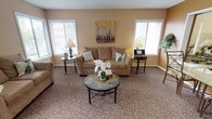 The Club Room at 702 W Casino Rd in Everett, Washington offers a comfortable and inviting community gathering space spread across two floors. The main level features a bright, carpeted lounge area furnished with plush sofas, upholstered accent chairs, and decorative side tables. Large windows fitted with white blinds allow generous natural light to fill the room, creating a warm and airy atmosphere complemented by soft, neutral tones throughout. A tiled entryway with French doors leads into the seating area, illuminated by an elegant chandelier that adds a refined touch. The upper level showcases dramatic vaulted ceilings with exposed beams, adding architectural character and a sense of openness. A chandelier anchors the space, while a wall-mounted flat-screen TV makes it ideal for group viewing. Cozy seating arrangements, including dark upholstered sofas and accent chairs, give this level a relaxed cinematic lounge feel, accented by decorative "Cinema" signage and greenery. Throughout both floors, patterned carpeting, warm peach and beige walls, and thoughtful décor create a cohesive, welcoming ambiance suitable for residents to gather, socialize, and unwind.