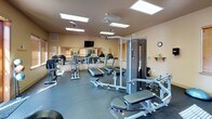 Located at 2711 East Adirondack Lane in Spokane, Washington, this well-equipped fitness center is designed to support a comprehensive workout experience on a single level. The space is bathed in natural light streaming through multiple windows fitted with wood-trimmed blinds, complemented by recessed panel ceiling lights that ensure bright, even illumination throughout.

The main workout area is generously proportioned, featuring rubber flooring ideal for both cardio and strength training activities. Cardio equipment includes multiple treadmills and elliptical machines, along with a recumbent stationary bike positioned near a glass door that opens directly to the outdoors, offering views of the surrounding landscaped grounds. A wall-mounted flat-screen television provides entertainment during workouts, and a large wall mirror expands the sense of space while allowing users to monitor their form.

The strength training section is equally well-appointed, housing a multi-station cable weight machine, an adjustable weight bench, a full rack of dumbbells in graduated sizes, and exercise balls. Floor mats are available for stretching and floor exercises. A bulletin board and informational displays are mounted on the walls, adding a functional touch. The overall ambiance is clean, practical, and welcoming, making this fitness center a comprehensive facility for residents.