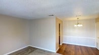 This single-floor residence offers a straightforward and functional layout designed for comfortable everyday living. The interior is finished with neutral, warm-toned walls that provide a versatile backdrop adaptable to a variety of décor styles. The living area features wall-to-wall carpeting underfoot, contributing to a cozy and quiet atmosphere, while adjacent spaces transition to rich, dark hardwood-style flooring that adds warmth and visual contrast throughout the home. Ceilings throughout are finished with a classic textured popcorn treatment, and ceiling-mounted HVAC vents ensure consistent climate control. A partial wall divides the main living space from a connecting area, hinting at a closet or hallway beyond, and a wall-mounted sconce light fixture provides ambient accent lighting in the adjacent room. Clean white baseboards frame each room, adding a crisp, finished look to the neutral palette. The open sightlines between rooms create a sense of flow and connectivity across the single floor. Overall, the space presents a clean, move-in ready canvas well-suited for personalization.