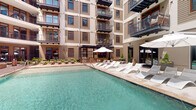 This outdoor amenity space is set within a multi-story residential complex, presenting a well-designed courtyard pool area that serves as the centerpiece of the community. The sparkling turquoise pool is generously proportioned and is flanked by a wide, paved pool deck finished in warm-toned pavers and natural wood-look decking on one side, creating a visually dynamic surround.

The pool deck is furnished with multiple loungers — including both cushioned green sling chairs and white chaise lounges — positioned to maximize sun exposure. Large market umbrellas in white are strategically placed throughout, providing shaded retreats. Additional seating clusters with bistro-style tables and chairs offer spaces for casual gatherings.

The courtyard is enclosed on multiple sides by the building's facade, which features horizontal lap siding in warm beige tones, wood-trimmed windows, and private balconies with metal railings that overlook the pool below. Raised planter beds filled with lush greenery and seasonal flowers add natural warmth to the space.

A dedicated lounge and grilling area with a covered pavilion anchors one end of the courtyard, while mature trees visible beyond the perimeter walls frame the sky above, enhancing the open-air atmosphere. The overall ambiance is resort-like, combining urban architecture with comfortable outdoor living.