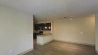 This single-floor space features an open-concept layout that seamlessly connects the kitchen and living areas, creating a spacious and functional environment. The kitchen is outfitted with rich, dark cabinetry that provides a striking contrast against the light-toned walls and countertops. A peninsula with a granite or stone-style countertop extends into the living area, offering additional workspace and casual seating opportunities. Pendant lights hang above the peninsula, adding focused illumination and a contemporary design element. Stainless steel appliances, including a microwave and range, are integrated into the kitchen's dark cabinetry for a cohesive look. Track lighting runs along the ceiling of the living area, providing adjustable, even lighting throughout the space. The flooring throughout is a warm, wood-style hardwood or laminate, adding texture and continuity across the open floor plan. Neutral, light-beige walls complement the darker kitchen finishes, contributing to a balanced and modern aesthetic. The overall ambiance is clean, contemporary, and well-lit, making the space feel welcoming and versatile.