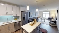 This single-floor residence offers a thoughtfully designed open-concept layout that seamlessly connects the kitchen, dining, and living areas into one cohesive living space. The kitchen is outfitted with sleek white cabinetry, a light-toned quartz countertop island, stainless steel appliances including a French door refrigerator and dishwasher, and a soft green glass tile backsplash that adds a subtle pop of color. Pendant lighting hangs above the island, providing both functional illumination and a stylish design accent. The dining area sits just beyond the kitchen, offering a natural transition into the open living space. The living room is bathed in natural light from a large window, creating a bright and welcoming atmosphere. Walls are painted in a cool, calming light blue-grey tone that complements the contemporary aesthetic throughout. Light hardwood-style flooring flows continuously across the space, enhancing the sense of openness and cohesion. The overall ambiance is clean, modern, and inviting, with a neutral palette accented by warm decorative touches that make the space feel both stylish and comfortable.