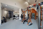 Located at 5751 Wood Meadow Way in Centreville, Virginia, this expansive fitness center offers a comprehensive workout environment spread across a generously sized single-floor layout. The space is thoughtfully divided into distinct functional zones, each designed to support a wide range of fitness activities.

The strength training area is a standout, anchored by striking orange-and-black HOIST multi-functional training rigs, TRX suspension systems, a full rack of dumbbells, and adjustable benches. This zone is adorned with bold, geometric murals in orange, navy, and cream tones, set against large wall mirrors that amplify the sense of space. The rubber-floored area provides excellent traction and durability for heavy lifting.

Transitioning into the cardio wing, the space opens up with high ceilings, ceiling fans, and multiple windows that invite natural light and outdoor views. A long row of TRUE-brand treadmills lines one wall, complemented by stationary bikes, ellipticals, and stair climbers, all arranged for easy access and clear sightlines.

Additional selectorized strength machines and cable systems occupy a dedicated section, rounding out the facility's full-body training capabilities. The flooring throughout the cardio zone features a striped commercial carpet with blue accent lines, lending a clean, modern aesthetic. Recessed lighting and ceiling fans provide a comfortable environment, making this fitness center a well-appointed destination for all levels of fitness enthusiasts.