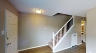This two-storey space is characterized by a clean, straightforward design that emphasizes functionality and comfort. The main level showcases warm hardwood-style flooring that runs seamlessly throughout, complemented by neutral grey walls and crisp white baseboards that lend a modern, cohesive aesthetic to the interior.

A prominently positioned staircase connects the two floors, featuring carpeted treads for added comfort underfoot, alongside a classic white balustrade with neatly spaced spindles and a sturdy newel post. The staircase design adds a traditional architectural touch while maintaining a clean, timeless look.

The ceiling features a textured finish and is fitted with a flush-mount light fixture that provides warm, ambient illumination to the space. The angled ceiling line above the staircase adds visual interest and a sense of dynamic height to the interior.

Overall, the space presents a well-maintained, neutral palette that offers a versatile canvas, making it well-suited to a variety of furnishing styles and personal tastes. The combination of durable flooring, classic staircase detailing, and consistent finishes throughout contributes to a welcoming and practical living environment.