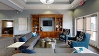 The Orchard Meadows leasing office is a thoughtfully designed single-story space that blends professional functionality with a comfortable, welcoming atmosphere. The interior is finished in cool slate-blue tones with crisp white crown molding and tray ceiling details, lending the space an elevated, polished character throughout.

The main lounge area serves as the heart of the office, anchored by a substantial warm-toned wood built-in wall unit housing a flat-screen television, open display shelving, and closed cabinetry. Seating arrangements include a generous gray tufted sofa, coordinating accent chairs upholstered in bold chevron fabric, and metallic drum-style coffee tables that add a contemporary touch. Recessed lighting and a statement pendant fixture illuminate the space evenly.

A welcoming reception zone near the entrance features glazed doors that draw in natural light, alongside a small refreshment station for visitors. Adjacent to the lounge, a dedicated leasing office workspace is equipped with a wood desk, guest seating, and a multifunction printer, with the Orchard Meadows branding prominently displayed on the wall. Carpeted flooring runs throughout, complementing the cohesive, professional ambiance of the entire space.