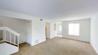 This two-floor space presents a clean, open layout with a design centered on functionality and simplicity. The main living area is generously proportioned, featuring wall-to-wall neutral beige carpeting that extends seamlessly into adjoining rooms, creating a cohesive flow throughout the interior. Bright white walls and smooth ceilings contribute to an airy, light-filled atmosphere.

A partial dividing wall separates the main living space from a secondary area, which is visible through a central opening. Built-in shelving is recessed into this dividing wall, offering practical storage or display space. The secondary room benefits from a large window that allows natural light to filter through, brightening both areas simultaneously.

To one side of the main living area, a bifold-door closet provides convenient storage. HVAC vents are positioned in both the floor and ceiling, ensuring efficient climate control throughout. Electrical outlets are evenly distributed along the walls.

The overall ambiance is neutral and move-in ready, with a straightforward, practical aesthetic that provides a versatile canvas for a variety of interior arrangements and styles across both floors.