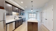 Located at 1310 Frost Ave in Maplewood, MN, this well-designed one-bedroom, one-bathroom unit offers a thoughtful layout across a single floor. The open-concept kitchen and living area serves as the heart of the space, featuring rich dark-toned cabinetry, stainless steel appliances including a refrigerator, range, microwave, and dishwasher, complemented by a tile backsplash and light quartz countertops. A central kitchen island with pendant lighting above provides additional prep space and casual seating, while light hardwood-style flooring flows throughout the main living area. Large windows at the end of the living space invite generous natural light and offer views of the surrounding outdoor scenery. The bedroom is carpeted for comfort and includes its own closet, with direct access to both the bathroom and the main living area. A second closet adds extra storage convenience. The bathroom features a full tub-shower combo, a vanity with dark wood cabinetry, and speckled tile flooring with matte black hardware accents throughout. A dedicated in-unit laundry area rounds out the functional amenities. The overall aesthetic is clean, contemporary, and move-in ready.