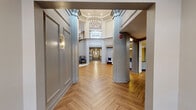 Located at 1551 Johnson Ferry Rd in Marietta, Georgia, this sophisticated common area space presents an impressive blend of classical architectural design and refined modern finishes. The grand foyer serves as the centerpiece, showcasing soaring two-story ceilings, a sweeping staircase with elegant wood-and-iron railings, and a striking chandelier that casts warm light throughout. Decorative wainscoting and raised panel wall moldings in soft gray tones lend a polished, upscale character to the space. Herringbone-patterned hardwood flooring flows seamlessly throughout, connecting the foyer to the extended hallways and gathering areas. A seating vignette beneath the staircase, appointed with wingback chairs and a decorative side table, offers an inviting resting spot. The adjacent gathering room features an ornate embossed tin ceiling with gold accents, herringbone flooring, a large marble-top communal table, pendant lighting in brushed brass, and a built-in cabinetry bar area with navy cabinetry and marble countertops. Curated wall art and carefully placed décor complete the refined aesthetic. Double glass entry doors framed by tall transom windows allow natural light to fill the entrance corridor, reinforcing the bright, welcoming ambiance of this thoughtfully designed community space.