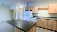 This single-floor residential space is designed with functionality and simplicity in mind, offering a clean, modern aesthetic throughout. The kitchen serves as the heart of the unit, showcasing light wood cabinetry that extends from the base to the upper walls, providing ample storage for everyday essentials. A dark-topped central island anchors the kitchen, offering additional prep space and a casual gathering spot. The kitchen is well-equipped, featuring a full-size white refrigerator, a dishwasher, and a double-basin sink with a modern faucet, all set against dark laminate countertops that provide a subtle contrast to the light cabinetry. Light wood-toned flooring runs throughout, contributing to a warm and cohesive feel. Recessed ceiling lighting ensures the space is well-lit, enhancing the open and airy atmosphere. A solid entry door is visible from the kitchen area, offering a sense of the unit's layout and accessibility. The overall design is practical and contemporary, making efficient use of the available space while maintaining a tidy, welcoming ambiance suited for comfortable day-to-day living.