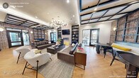 This thoughtfully designed single-floor space masterfully blends industrial character with contemporary sophistication across a generous footprint. The open-plan layout flows seamlessly between a lounge area and a fully equipped kitchen and dining zone, creating a highly functional and visually cohesive environment.

The lounge area commands attention with its dramatic coffered ceiling accented by dark steel beams, a striking mid-century modern chandelier, and a wall-mounted fireplace flanked by floor-to-ceiling built-in bookshelves. Rich leather sofas, curated décor, and warm hardwood flooring contribute to an inviting, upscale atmosphere. Glass doors open directly to an outdoor patio, allowing natural light to pour in and offering views of the surrounding landscape.

The kitchen features a long, waterfall-edge marble island with leather bar stools, complemented by sleek gray cabinetry, open shelving with under-cabinet lighting, and gold-toned dome pendant lights. The space is equipped with stainless steel appliances, making it well-suited for gatherings and events.

A wide corridor bordered by exposed brick on one side and steel-framed glass partitions on the other connects the main areas. The brick walls, present throughout, lend an authentic industrial warmth that anchors the space's design identity. Private rooms accessible via glass-panel doors offer flexibility for focused use or meetings.
