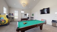 Located at 7027 Park Mill Dr in Plain City, Ohio, this single-story community amenity facility is thoughtfully designed to accommodate both recreation and formal gatherings. The centerpiece of the space is a spacious game room highlighted by soaring vaulted ceilings and an elegant chandelier overhead. This area is outfitted with a full-size billiard table, a foosball table, a classic arcade game, and a wall-mounted flat-screen TV, all set on durable patterned carpet. A large Connect Four game adds a playful touch, and multiple windows allow natural light to pour in while offering views of the surrounding area. A teal accent wall adds a vibrant pop of color and visual interest to this lively zone. Adjacent to the game room is a dedicated card and poker room, furnished with a professional oval poker table and black leather chairs. A wall-mounted TV makes it equally suited for watching sports or hosting casual gatherings. The space also includes a formal conference or meeting room, appointed with a long rectangular table, upholstered chairs, and generous windows fitted with white blinds that provide an abundance of natural light and exterior views. Neutral tones, clean white walls, and consistent carpeting throughout create a cohesive and welcoming environment across all areas.