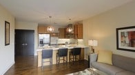 This single-floor residence showcases a thoughtfully designed open-concept layout that integrates the kitchen and living area into one cohesive and welcoming environment. The kitchen is a standout feature, outfitted with rich warm-toned wood cabinetry that extends to the upper walls, providing generous storage while adding natural warmth to the space. Stainless steel appliances, including a French door refrigerator, lend a contemporary edge, while a sleek light-colored countertop extends outward to form a peninsula-style breakfast bar. Three dark upholstered bar stools line the bar, creating a casual dining and gathering spot. Above, two globe-style pendant lights hang at varying heights, casting a warm ambient glow and serving as a focal point of the kitchen's design. A textured tile backsplash adds visual interest and dimension behind the cooking area. Dark hardwood flooring runs throughout, grounding the space with depth and elegance and creating a striking contrast against the lighter cabinetry and countertops. The adjacent living area transitions seamlessly from the kitchen, furnished with a comfortable sofa and complementary accent pieces. Together, the spaces offer a refined, modern aesthetic that is both functional and inviting.