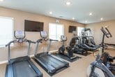 This fitness center offers a comprehensive workout environment designed to support a variety of exercise routines. The single-floor space is finished with neutral-toned walls and low-pile carpeting, creating a clean and functional atmosphere throughout. Recessed lighting illuminates the room evenly, complemented by natural light admitted through multiple windows fitted with blinds.

The cardio section is prominently stocked with True-brand equipment, including treadmills, elliptical trainers, and a stationary bike, each positioned on protective floor mats to preserve the flooring. Multiple wall-mounted flat-screen televisions are positioned throughout the space, allowing users to stay entertained during cardio sessions.

The strength training area features a multi-station cable machine, an adjustable weight bench, and a well-organized dumbbell rack holding a wide range of free weights. A wall-mounted fan provides additional air circulation, and a yoga mat hung on the wall adds versatility for stretching or floor exercises. A small storage unit and sanitation station are also present, supporting cleanliness and convenience. The overall layout is open and thoughtfully arranged to allow comfortable movement between stations.