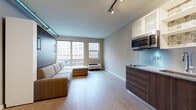 This single-floor studio with one bathroom offers a thoughtfully designed open-concept layout that maximizes functionality without sacrificing comfort. The main living area is bright and airy, featuring large windows that frame views of the surrounding urban cityscape and allow natural light to fill the space. Light hardwood-style flooring runs throughout, complementing the neutral tones of the walls and creating a cohesive, modern aesthetic.

A standout feature is the wall-mounted Murphy bed system, which folds away to reveal an L-shaped sectional sofa, seamlessly transforming the sleeping area into a spacious living room. Built-in open shelving alongside the Murphy bed provides convenient storage and display space. An exposed ductwork detail along the ceiling adds an industrial touch, while track lighting above ensures the space is well-illuminated.

The kitchenette is efficiently designed with dark wood cabinetry, granite-style countertops, a stainless steel refrigerator, built-in microwave, flat cooktop, and a sink — all compactly arranged to optimize workflow. White upper cabinets with glass-front panels add a light contrast to the lower cabinetry. A dedicated workspace with a desk rounds out the room.

The bathroom is clean and functional, featuring a combination tub and shower with white tile surround, chrome fixtures, and a towel rack, maintaining the space's consistent, understated style.