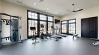 Located at 37 Foxgrove Dr in Dawsonville, Georgia, this dedicated fitness center offers a thoughtfully designed exercise space built to support a full range of workout routines. The single-room layout makes efficient use of its footprint, providing ample open floor space covered in durable, speckled rubber flooring ideal for high-impact activity.

Large, black-framed floor-to-ceiling windows line the main wall, flooding the space with natural light and offering views of the surrounding neighborhood. The bright, neutral-toned walls are complemented by a warm amber accent wall that anchors the strength training zone, adding visual depth and energy to the room. Recessed lighting and a modern ceiling fan with sleek black blades ensure the space remains well-lit and comfortably ventilated during workouts.

The cardio section is outfitted with treadmills, an elliptical, and a stationary bike, all positioned to face the expansive windows. A wall-mounted flat-screen TV is conveniently located nearby. The strength area features a multi-function power rack with weight plates, a dedicated dumbbell rack with a full range of free weights, medicine balls, resistance bands, stability balls, and a adjustable weight bench. Black double doors provide entry and exit access. The overall ambiance is clean, modern, and highly functional.