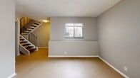 This two-story space is designed with a clean, neutral aesthetic, utilizing soft gray tones, white trim, and light flooring throughout to create a cohesive and modern feel. The main level features an open living area with light wood-style flooring, a window with blinds, and a sliding glass door that invites natural light. A staircase with black metal railings connects the two floors. The kitchen is efficiently laid out in an L-shape, equipped with gray cabinetry, granite countertops, a stone tile backsplash, and stainless steel appliances including a range, dishwasher, and refrigerator. A half bathroom on the main level features a granite-topped vanity, an undermount sink with oil-rubbed bronze fixtures, and a round black-framed mirror. Upstairs, the bedrooms feature vaulted ceilings with textured finishes, light flooring, and ample closet space. One bedroom includes a wide horizontal window with frosted glass for privacy, while another offers a standard window with blinds and an open closet. A full bathroom upstairs mirrors the same granite vanity style and includes a tiled tub-shower combination, maintaining design consistency throughout the space.