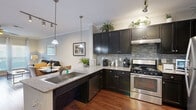 This single-floor residence offers a thoughtfully designed open-concept layout that seamlessly connects the kitchen, dining, and living areas. Rich hardwood flooring and crown molding run throughout the main living spaces, contributing to a polished and cohesive aesthetic.

The kitchen is a standout feature, outfitted with dark espresso cabinetry extending to the ceiling, light granite countertops, and a mosaic tile backsplash. Stainless steel appliances — including a gas range, dishwasher, and refrigerator — complement the space, while pendant lighting over the peninsula adds both function and style. Track lighting provides additional illumination.

Adjacent to the kitchen, the dining area accommodates a full table and chairs and flows naturally into the living room, which features a ceiling fan, large windows with abundant natural light, and access to an outdoor area via glass doors.

A hallway connects the living spaces to the private quarters, where the bedroom offers plush carpeting, generous natural light from multiple windows, a ceiling fan, and direct access to a walk-in closet. The neutral, light gray wall palette used throughout creates a calm and inviting atmosphere across the entire residence.