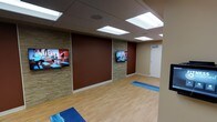 This single-floor fitness studio is thoughtfully designed to support a variety of exercise and wellness activities. The room features warm hardwood-style flooring that provides a comfortable and durable surface ideal for yoga, stretching, and low-impact workouts. Exercise mats are laid out on the floor, ready for use.

The walls present a striking combination of rich terracotta-toned painted panels and natural stacked stone accent sections, creating an energizing yet grounded aesthetic. Multiple flat-screen televisions are mounted along the walls, allowing participants to follow guided workout or yoga video sessions with ease from various positions throughout the room.

The ceiling is fitted with a drop-tile system and flush rectangular light panels that provide bright, even illumination across the entire space, ensuring a well-lit environment for all activities. A standard door on one end of the room provides access, keeping the layout clean and functional.

Overall, the studio strikes a balance between practicality and visual appeal, offering a welcoming environment for fitness enthusiasts. The combination of warm tones, quality materials, and media-enabled walls makes this space well-suited for both guided class formats and independent exercise sessions.