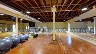 This single-floor fitness studio offers a well-appointed environment designed for group exercise and indoor cycling. The space is defined by its striking exposed wood-beam ceiling, which gives the room an industrial-chic character, complemented by recessed lighting and visible ductwork that add to the open, loft-like aesthetic. The rich hardwood flooring throughout provides both durability and warmth, reflecting the overhead lights for a vibrant, energetic atmosphere.

Along one wall, a full row of stationary cycling bikes is neatly arranged, mirrored by large wall-to-wall mirrors that amplify the sense of space and allow participants to monitor their form during workouts. A mounted flat-screen television is positioned at the front of the room, supporting instructor-led sessions and class programming. A vertical storage rack holds an organized array of colorful hand weights, making free-weight exercises easily accessible.

Stability balls are also present on the studio floor, indicating the space accommodates a variety of fitness modalities beyond cycling. A central structural column adds to the architectural character without impeding functionality. The overall ambiance is energetic yet focused, making this studio well-suited for cycling classes, circuit training, and other group fitness formats.