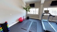 Located at 2355 Michigan Ave in Pensacola, Florida, this fitness space offers a practical and well-appointed environment designed to support a variety of workout routines. The single-floor layout is thoughtfully organized to maximize usability while maintaining an open, uncluttered feel. The room features rubber flooring ideal for exercise, providing both durability and comfort underfoot. Natural light streams in through a sliding window, brightening the space and creating an inviting atmosphere. A wall-mounted flat-screen TV is positioned at an optimal viewing angle, allowing users to follow workout content or stay entertained during cardio sessions. The space is equipped with a treadmill for cardiovascular training, along with exercise mats, a stability ball, and a step trainer for strength and flexibility work. A potted indoor tree adds a touch of greenery, contributing to a calm and refreshing ambiance. The neutral-toned walls and clean lines of the room reflect a modern, no-frills aesthetic focused entirely on function. Whether used for cardio, stretching, or core training, this fitness space provides a comfortable and well-equipped environment suited for daily exercise routines.