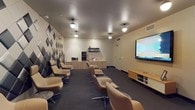 This purpose-built screening room offers a refined, theater-like environment designed for an immersive viewing experience. The space is anchored by a large flat-panel display screen mounted on the front wall, paired with a low-profile media console that houses the necessary equipment for seamless presentations or screenings. Rows of comfortable tan leather swivel chairs are arranged in a gentle arc, providing clear sightlines to the screen from every seat and lending the room a polished, professional atmosphere.

The interior design balances functionality with aesthetic appeal. A striking geometric accent wall, featuring a diamond pattern in contrasting dark and light tones, serves as a bold focal point and also provides acoustic treatment to enhance sound quality within the room. The remaining walls are finished in a warm neutral tone, contributing to a calm and focused ambiance.

Along the back wall, a well-appointed refreshment station includes cabinetry, a microwave, a beverage refrigerator, and open shelving — ensuring guests have everything they need for an extended viewing session. Overhead, a series of pendant and flush-mount ceiling lights provide even, adjustable illumination throughout the space. Dark carpeting grounds the room and further aids in sound absorption, completing the cinematic feel of this thoughtfully designed screening environment.