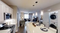 This single-floor residence offers a thoughtfully designed open-concept layout that seamlessly connects the kitchen, dining, and living areas into one cohesive, inviting space. The kitchen is a standout feature, showcasing white cabinetry topped with light-toned quartz countertops that extend into a generous island with seating. Overhead, a trio of sleek glass pendant lights adds a modern, refined touch. A stainless steel range anchors the cooking area, completing the kitchen's contemporary appeal. The dining space flows naturally from the island, comfortably accommodating a table and chairs for everyday meals or entertaining. Beyond, the living room is bathed in natural light from a large window dressed with elegant draping curtains. Rich, dark hardwood-style flooring runs throughout, grounding the space with warmth and sophistication. Neutral gray walls create a calm, versatile backdrop that complements the modern furnishings. A ceiling fan adds both style and function to the living area. A glimpse through an open doorway reveals a bedroom, suggesting a well-connected and practical floor plan. Overall, the space exudes a polished, contemporary ambiance suited for comfortable everyday living.