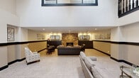 Located at 10134 W Mohawk Ln in Peoria, Arizona, this impressive communal lounge space makes an immediate statement with its soaring double-height ceilings and an abundance of natural light filtering in through large windows and skylights above. A dramatic black-railed staircase anchors the room, ascending elegantly beneath a striking candelabra-style chandelier that adds a touch of sophistication to the open layout.

The main lounge area is thoughtfully organized into distinct yet connected zones. A comfortable seating arrangement, centered around a glass-topped coffee table, provides an inviting space for casual conversation, while a dedicated dining nook with a round wooden table and upholstered chairs offers a more intimate setting. A media console with a wall-mounted flat-screen television and a classic accent chair complete the entertainment area.

Toward the rear, a standout stone fireplace serves as a dramatic focal point, flanked by dark wood cabinetry and decorative mirrors that reflect warm ambient lighting. Additional seating is arranged around this area, creating a cozy retreat within the larger space. A consistent design language — warm neutral tones, dark wood trim accents running along the walls, and plush carpeting — ties all zones together cohesively, resulting in a welcoming and well-appointed communal environment.