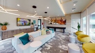 This stylish single-floor community clubroom is designed for socializing and relaxation, offering a thoughtfully curated mix of leisure and entertainment amenities. The main lounge area centers around a large, plush modular sofa accented with colorful teal, yellow, and multicolored throw pillows, complemented by rounded side tables and modern cylindrical ottomans. Striking geometric pendant light fixtures hang from the ceiling, adding a contemporary flair alongside recessed lighting throughout. A bold teal accent wall anchors one side of the space, where a wall-mounted flat-screen TV provides a media focal point. Adjacent to the lounge, a casual game table with wooden chairs offers a space for board games beneath another mounted television. A dedicated recreation area features a full shuffleboard table set against a textured stone accent wall adorned with vibrant large-scale artwork. Built-in cabinetry along one wall provides practical storage and a print station. Floor-to-ceiling windows and sliding glass doors flood the interior with natural light while framing views of an outdoor pool and patio area, creating a seamless indoor-outdoor connection. Patterned carpeting with an abstract design ties the space together, contributing to its lively yet sophisticated ambiance.