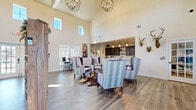 This single-story community clubhouse offers an impressive array of shared amenities designed for comfort, activity, and convenience. The centerpiece is a grand, open-concept great room distinguished by soaring vaulted ceilings, statement orb chandeliers, and an abundance of natural light streaming through clerestory windows and large exterior windows. Rich hardwood-style flooring runs throughout, anchoring a stylish seating arrangement of upholstered wingback chairs accented by rustic décor, including mounted antler and animal head wall pieces that lend a warm, lodge-inspired character.

Adjacent to the great room is a well-appointed communal kitchen featuring dark espresso cabinetry, sweeping granite countertops, a large central island with bar seating, beverage refrigerators, and pendant lighting — ideal for social gatherings. A secondary kitchenette with gray cabinetry provides additional convenience.

The fitness center is generously sized, outfitted with cardio machines including treadmills, an elliptical, a stair climber, and a stationary bike, along with strength equipment and free weights, all reflected by full-length mirrored walls and lit by natural light from multiple windows.

A dedicated business center offers built-in workstations with multiple computer monitors. The covered outdoor patio, framed by a wood-planked ceiling and brick exterior, provides a relaxed setting overlooking a landscaped courtyard with additional seating.
