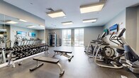 Located at 28 Southfield Ave in Stamford, Connecticut, this modern fitness center offers a comprehensive and thoughtfully arranged workout facility on a single floor. The space is designed to accommodate a wide range of exercise routines, from cardiovascular training to strength conditioning.

The cardio zone features a row of treadmills and elliptical trainers, each equipped with individual screens for entertainment during workouts. Large wall-mounted flat-screen TVs add to the engaging atmosphere. Floor-to-ceiling windows along one wall allow natural light to fill the space and provide views of the surrounding exterior.

The strength training area is well-stocked with a full rack of dumbbells spanning a wide weight range, adjustable workout benches, and a multi-station cable machine. Exercise and stability balls are neatly organized on wall-mounted racks, supporting functional fitness and stretching routines.

Full-length mirrors line multiple walls, visually expanding the space and allowing users to monitor their form. The flooring throughout is durable rubberized gym material, providing both safety and comfort underfoot. Recessed and panel flush lighting in the ceiling ensures bright, even illumination across the entire facility.

The overall ambiance is clean, modern, and professional, with a neutral gray and white color palette that creates an energizing yet focused environment suited for a variety of fitness goals.