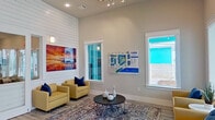 This single-story clubhouse for the Tide community is designed with a lively coastal aesthetic, immediately recognizable by its bold turquoise exterior with crisp white trim. The entrance opens through glass-paneled French doors into a bright, airy lobby featuring wood-look tile flooring, shiplap accent walls, and soaring ceilings. A welcoming lounge area near the entry is furnished with yellow armchairs, a round coffee table, and a patterned area rug, complemented by vibrant coastal artwork and a community map display. Glass-paned double doors lead into a spacious social room bathed in natural light from large windows on all sides, featuring an elegant chandelier and an arrangement of navy blue seating — ideal for gatherings. A separate office or leasing area with sliding glass doors provides a private space for resident services. The fitness center is generously sized, offering abundant natural light from transom and full-size windows, and is equipped with treadmills, a stair climber, a cable machine, free weights, and a dedicated studio area with spin bikes, a rowing machine, and hanging pendant fixtures. The covered outdoor patio, painted in the signature turquoise, provides a shaded retreat with striped lounge seating overlooking the surrounding community and pool area. A modern kitchen rounds out the amenities, featuring granite countertops, white cabinetry, stainless steel appliances, and wood-look tile flooring.