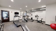 Located at 719 Virginia Ave in Indianapolis, IN, this fitness center offers a well-equipped exercise room designed to support a well-rounded workout routine. The space features a clean, modern aesthetic with crisp white walls, recessed lighting, and durable carpet tile flooring that provides both comfort and traction underfoot. Exposed ductwork along the ceiling adds an industrial touch to the otherwise streamlined interior.

The room is outfitted with a comprehensive selection of cardio equipment, including multiple treadmills from True and Spirit, an elliptical trainer, and a stationary bike, giving users a variety of options for cardiovascular exercise. A wall-mounted flat-screen television accompanies the cardio machines, enhancing the workout experience. For strength training, the space includes a rack of dumbbells in graduated weights, a adjustable workout bench, wall-mounted pull-up bars, and a stability ball with weight plates. A large wall mirror expands the visual depth of the room and assists with form checks during exercise.

A posted fitness area rules sign keeps the environment organized and user-friendly. The room benefits from recessed ceiling lights that ensure consistent, bright illumination throughout. The overall ambiance is functional and focused, offering residents or guests a practical and well-maintained space to maintain their fitness routines.