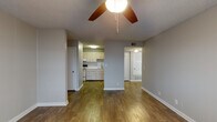 This single-floor unit is designed with a practical, open layout that connects the main living areas seamlessly. The living room is a comfortable, open space finished with warm wood-look flooring and neutral gray walls, with a ceiling fan providing both light and airflow. A textured popcorn ceiling is consistent throughout the unit. The living area flows naturally toward the kitchen, which is separated by a partial wall divider, creating a semi-open feel while maintaining functional distinction between spaces.

The kitchen is efficiently arranged along a single wall, featuring cream-toned cabinetry, a stainless steel sink with a modern faucet, a countertop range, and ample upper and lower cabinet storage. Recessed under-cabinet lighting adds functionality to the workspace.

The bedroom is carpeted for comfort and includes a ceiling fan, as well as a sliding glass door that opens to an outdoor balcony, allowing natural light to filter in and providing an outdoor view. White vertical blinds offer privacy when needed.

The bathroom features a vanity with white cabinetry, light countertops, and a wall-mounted mirror, alongside a separate wet area housing a tiled bathtub and toilet. Wood-look flooring extends through the bathroom, maintaining visual consistency with the rest of the unit.