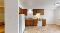 Located at 2450 Aurora Ave N in Seattle's Queen Anne neighborhood, this single-floor unit offers a straightforward and functional layout. The kitchen is equipped with warm oak-toned cabinetry spanning both upper and lower sections, providing ample storage space. White appliances include a coil-top electric range and a full-sized refrigerator, complemented by a stainless steel sink and light-toned laminate countertops. The kitchen floor features neutral stone-look tile, creating a clean and practical surface. Adjacent living and sleeping areas are finished with wall-to-wall carpet in a warm beige tone, contributing to a quiet and comfortable atmosphere. White walls throughout the unit keep the interior feeling open and bright. The bathroom includes a walk-in shower stall equipped with stainless steel grab bars for added accessibility, along with a toilet and a vanity with an integrated sink, all set on matching tile flooring. A dedicated closet area features sliding doors and built-in open shelving, offering organized storage. The unit's compact, elongated floor plan makes efficient use of the available space, with each area clearly defined and accessible from a central corridor.