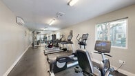 Located at 2850 Middlefield Road in Palo Alto, California, this fitness center offers a well-organized and fully equipped exercise space designed to support a comprehensive workout routine. The room features durable rubber flooring throughout, providing both safety and comfort underfoot. Fluorescent overhead lighting ensures the space is brightly and evenly lit, while multiple windows allow natural light to filter in, creating an open and energizing atmosphere. A full-length wall mirror along one side visually expands the room and allows users to monitor their form during exercise. The cardio section includes Life Fitness treadmills equipped with personal viewing screens, an elliptical trainer, a stationary upright bike, and a recumbent bike, offering a variety of low- and high-impact cardiovascular options. Strength training is supported by a multi-function cable machine and a rack of dumbbells spanning a range of weights, along with an adjustable weight bench. Climate control is provided by a wall-mounted split unit, ensuring comfort year-round. Decorative wall art adds a welcoming touch to the otherwise functional space. A glass-paneled door opens directly to an outdoor area, providing convenient access and additional ventilation.