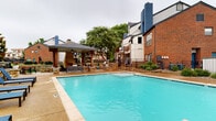 This inviting outdoor amenity space centers around a generously sized swimming pool with clear turquoise water, framed by a wide concrete pool deck designed for both relaxation and socializing. The surrounding brick architecture lends a classic, established character to the environment, complemented by mature trees that provide natural shade and a sense of privacy.

A standout feature is the covered cabana pavilion, supported by sturdy wooden posts and equipped with a ceiling fan for year-round comfort. The cabana includes a striking stone fireplace, comfortable lounge seating, and direct access to an outdoor kitchen with a built-in grill — making it an ideal gathering space for outdoor entertaining. Adjacent to the cabana, an outdoor dining area with umbrella-shaded tables and chairs offers a comfortable spot for poolside meals.

A dedicated sunbathing zone lined with blue lounge chairs provides ample space for relaxation. The pool area is accessed through a gated brick entryway, clearly marked with pool policies, ensuring a secure and well-managed environment. Lush landscaping with decorative stonework and greenery borders the perimeter, adding an attractive finishing touch to this comprehensive and well-maintained outdoor recreational space.