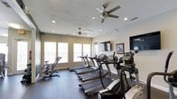 Located at 2000 Village Bluff Pl in Raleigh, NC, this fitness center offers a well-equipped, single-floor workout space designed to support an active lifestyle. The room is outfitted with a lineup of True-brand treadmills, stationary bikes, and elliptical machines, providing a comprehensive range of cardio options for residents or guests. The space is kept comfortable year-round with multiple ceiling fans and recessed lighting that create a bright, airy atmosphere. Natural light filters in through a row of large windows along one wall, fitted with horizontal blinds for adjustable illumination. Two wall-mounted flat-screen televisions are positioned for easy viewing during workouts, adding an element of entertainment to exercise sessions. The flooring is a durable dark material well-suited for high-traffic fitness use. The neutral-toned walls and clean layout give the space an organized, professional feel. Overall, this fitness center is thoughtfully designed to provide a functional and comfortable environment for a variety of cardio-focused workouts.