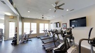 Located at 2000 Village Bluff Pl in Raleigh, NC, this fitness center offers a well-equipped, single-floor workout space designed to support an active lifestyle. The room is outfitted with a lineup of True-brand treadmills, stationary bikes, and elliptical machines, providing a comprehensive range of cardio options for residents or guests. The space is kept comfortable year-round with multiple ceiling fans and recessed lighting that create a bright, airy atmosphere. Natural light filters in through a row of large windows along one wall, fitted with horizontal blinds for adjustable illumination. Two wall-mounted flat-screen televisions are positioned for easy viewing during workouts, adding an element of entertainment to exercise sessions. The flooring is a durable dark material well-suited for high-traffic fitness use. The neutral-toned walls and clean layout give the space an organized, professional feel. Overall, this fitness center is thoughtfully designed to provide a functional and comfortable environment for a variety of cardio-focused workouts.