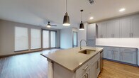 This single-floor residence showcases a thoughtfully designed open-concept layout that seamlessly connects the kitchen and living areas. The kitchen serves as a central hub, anchored by a large island with a light quartz countertop, an undermount sink with a brushed gold faucet, and seating overhang. Two statement pendant lights hang above the island, while gray shaker-style cabinetry lines the perimeter walls, complemented by white subway tile backsplash and a full suite of stainless steel appliances, including a refrigerator, range, and microwave. The adjoining living area benefits from natural light streaming through large windows and sliding glass doors, with a ceiling fan overhead. Wood-look flooring runs throughout the main living spaces, extending into the bedrooms. Each bedroom is well-proportioned with windows fitted with blinds and ceiling fans for comfort. The primary bathroom features an elongated dual-sink vanity with warm wood cabinetry, white quartz countertops, large-format white tile flooring, and access to a walk-in closet with open shelving. A secondary bathroom includes a tiled tub-shower combination with a built-in niche. A dedicated laundry area houses a full-size washer and dryer beneath a folding counter with overhead shelving.