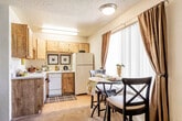 This single-floor residential unit offers a well-organized layout that maximizes its available space. The living area serves as the heart of the home, featuring carpeted flooring, a textured ceiling, and a semi-open design that connects seamlessly to the kitchen via a partial dividing wall. The living room provides ample space for comfortable seating arrangements and is brightened by interior lighting and neutral wall tones.

The kitchen is efficiently designed with warm wood cabinetry extending to the upper walls for generous storage, light-toned countertops, and hardwood-style flooring. It includes a full suite of appliances—a refrigerator, electric range, and dishwasher—all set within a compact yet functional galley-style layout illuminated by overhead fluorescent lighting.

The bedroom is a quiet, carpeted retreat with enough room for a full sleeping arrangement and additional furniture such as a desk or dresser. A textured ceiling continues throughout, and a doorway leads directly into the bathroom, offering convenient access.

The bathroom features a vanity with a granite-style countertop and wood cabinetry below, a wall-mounted mirror, and a tiled shower-tub combination. The clean, neutral finishes throughout the unit create a cohesive and practical living environment suited for everyday comfort.
