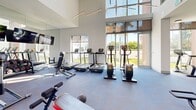 Located at 525 NE 7th St in Fort Lauderdale, FL, this well-appointed fitness center offers a spacious, single-floor exercise facility designed to support a complete workout experience. The space is defined by its striking architectural design, featuring soaring double-height ceilings in key areas and an abundance of floor-to-ceiling windows that flood the interior with natural light and provide open views of the surrounding lush greenery and outdoor landscape.

The exercise room is outfitted with a comprehensive suite of Technogym equipment, including multiple strength-training cable machines, a cable crossover station, adjustable benches, and free weights. The cardio section features treadmills, upright stationary bikes, and recumbent bikes, all positioned to take advantage of the natural light and outdoor views. A wall-mounted television adds to the functionality of the space, making it suitable for extended training sessions.

The interior finishes are clean and contemporary, with crisp white walls, durable exercise flooring, and recessed and pendant lighting that complement the abundant daylight. The overall ambiance is bright, open, and motivating, reflecting a modern aesthetic well-suited to an active Fort Lauderdale lifestyle. The generous footprint allows for comfortable spacing between equipment, ensuring an uncluttered and efficient workout environment.