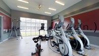 This single-story fitness center is designed to support a well-rounded cardio workout experience. The space is outfitted with a row of Matrix elliptical trainers and a spin bike, all positioned to maximize floor space while maintaining an open, accessible layout. The light wood-tone flooring adds warmth and is practical for a high-traffic exercise environment.

A large grid-pane window along one wall floods the room with natural light and provides views of the surrounding exterior, creating an airy atmosphere that enhances the workout experience. A ceiling fan overhead aids in air circulation, complementing the flush-mount fluorescent lighting that ensures consistent brightness throughout the space.

A bold red accent wall on one side adds a dynamic, energizing visual element that contrasts with the neutral gray walls elsewhere. The overall design balances functionality with an inviting aesthetic, making the space feel motivating and well-maintained. The layout allows for comfortable movement between equipment, and the clean, modern finishes throughout contribute to a polished and purpose-built fitness environment.