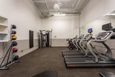 This fitness center, located in Atlanta, GA (30308), is a single-floor facility designed to support a comprehensive workout experience. The space features a clean, functional aesthetic with white walls accented by a two-tone lower border, carpet tile flooring, and flush-mounted ceiling lighting that keeps the environment bright and practical.

The cardio section is well-appointed with multiple Star Trac treadmills and a row of indoor cycling bikes equipped with interactive digital screens, ideal for both self-guided and virtual training sessions. A full-length wall mirror spans a significant portion of the room, visually expanding the space and allowing users to monitor their form during workouts.

Strength training is thoroughly covered with a multi-functional cable machine for resistance exercises, a dumbbell rack with a varied selection of free weights, and flat workout benches for versatile training. A dedicated area along one wall houses a medicine ball rack stocked with multiple weighted balls in various sizes, alongside a stability ball, offering options for functional fitness and core training.

The ceiling features exposed utility infrastructure, lending the space an industrial character while maintaining an overall clean and organized presentation. The layout is thoughtfully arranged to allow fluid movement between equipment zones, making this fitness center a practical and fully equipped resource for users of all fitness levels.